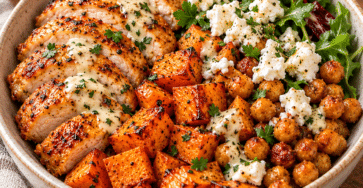 Warm chicken bowl with roasted sweet potato cubes, crispy chickpeas, crumbled feta, fresh salad greens, and lemon dressing served in a bowl.